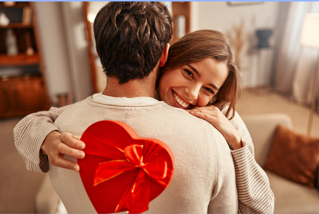 Woman hugging a man holding a heart-shaped gift box in a cozy living room.